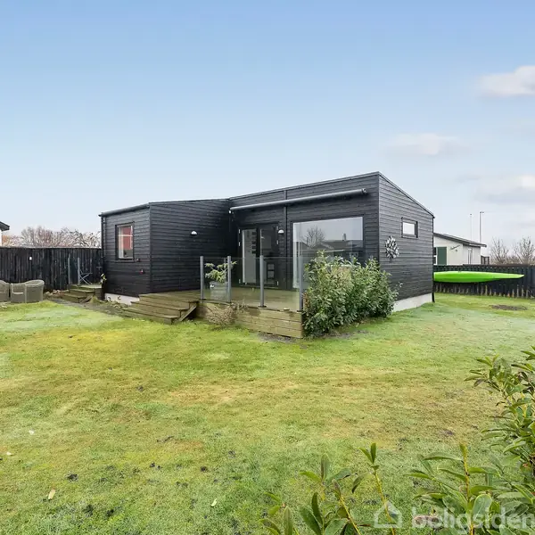 Et sort træhus står på en velholdt græsplæne med buske omkring. Huset har store vinduer og et terrasseområde med møbler. I baggrunden ses andre huse og en blå himmel.