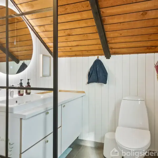 En gang med hvide trævægge og et rødt tæppe leder mod et rum med træloft. Til højre er et moderne badeværelse med en rund spejl og sort glasvæg.