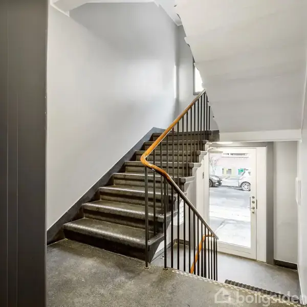 En grå trappe med gelænder fører opad i en opgang. Til højre er der en døråbning, hvorfra man kan se ud på en parkeringsplads med biler udenfor.