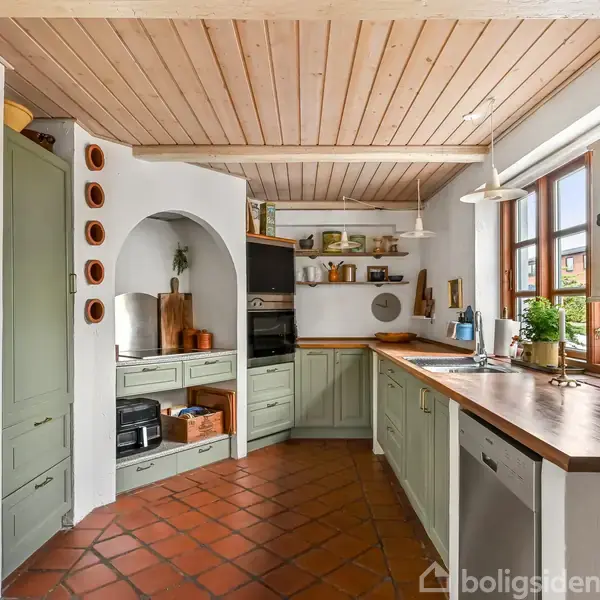 Et køkken med træloft og terracotta-flisegulv. Skabe og skuffer er malet i grønne nuancer. Der er en stor træbordplade ved vinduet, hvor planter står i solen.