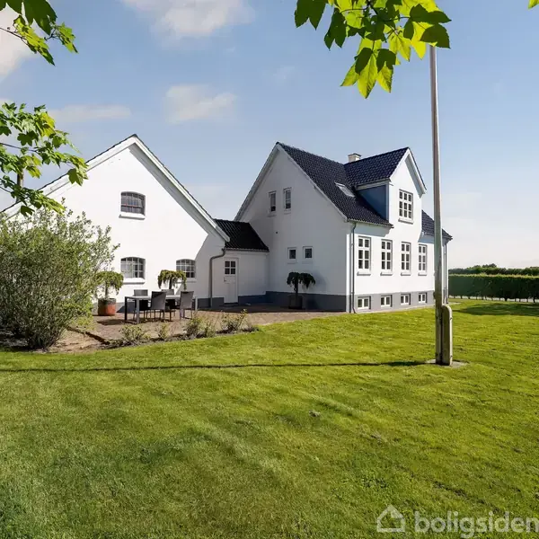 Hvidt, to-etagers hus står på en velplejet grøn græsplæne med en terrasse fyldt med havemøbler. Omkring huset er grønne træer og en åben himmel.