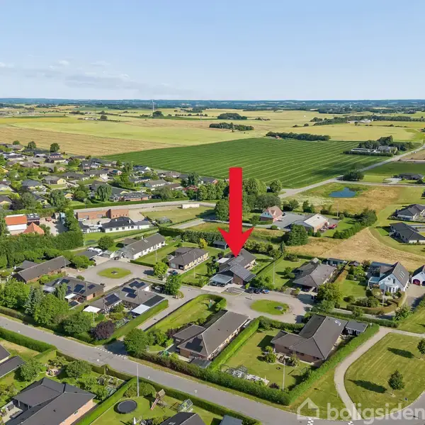 Rød pil peger på et hus i en forstadskvarter, omkranset af grønne områder og nabohuse. Stedet ligger i et åbent landskab med marker i baggrunden.