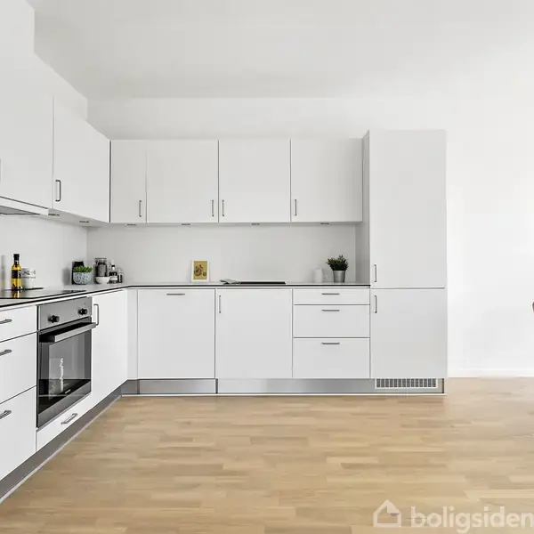 Hvidt køkken med indbyggede skabe og apparater, herunder en ovn og køleskab, i et rum med trægulv. Til højre står et spisebord med rosa stole og en grøn plante.