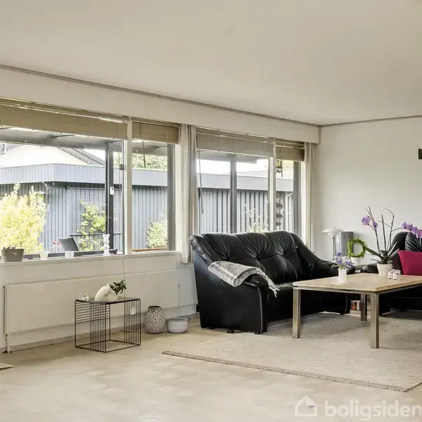 Sort sofa står i hjørnet af en lys stue, omgivet af store vinduer og en træbord med planter og dekorationer. Enkle gardiner hænger ned, og udenfor ses en terrasse med planter.