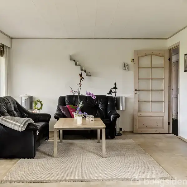 Sorte lædersofaer står omkring et træbord med blomster i en lys stue. Store vinduer bagved slipper naturligt lys ind. En trædør og et skab ses til højre.
