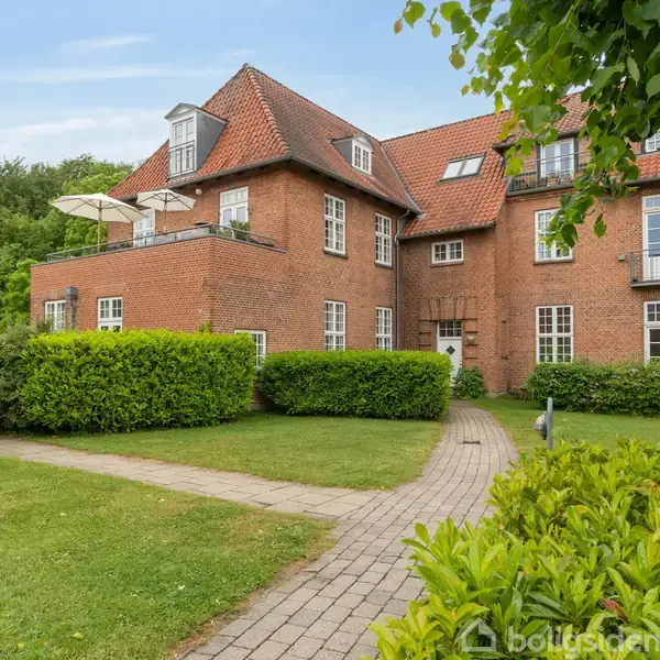 En murstensbygning med rødt tag står i en velplejet have. Bygningens facade har flere vinduer og altaner. En sti leder gennem græsset og buskene foran huset.