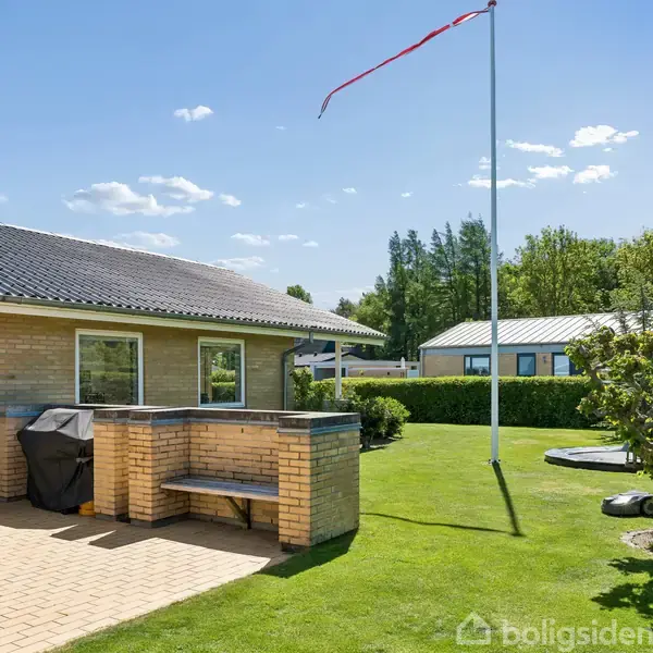 Hus med terrasse i solskinsvejr, omgivet af grøn græsplæne og buske. Spisebord og grill står på terrassen. Flag i baggrunden uden flaghejsning.
