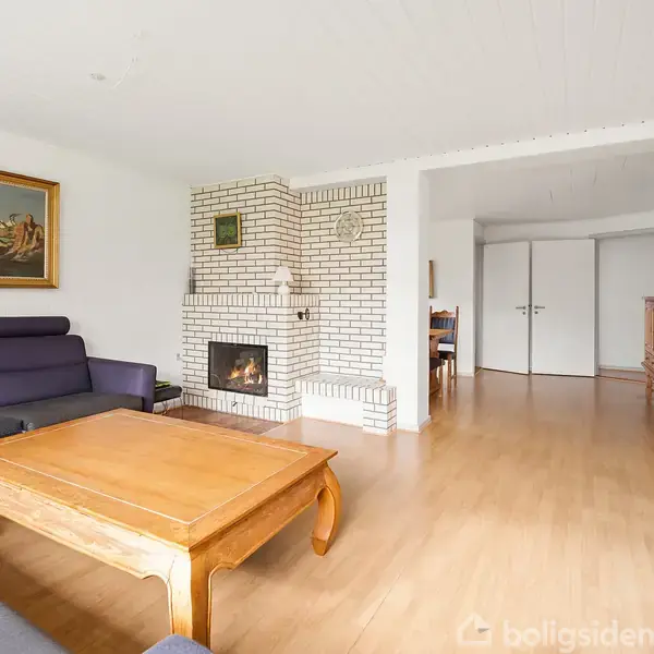 Sorte sofaer står omkring et træbord i en lys stue med trægulv, hvidmalede vægge og en hvid murstenspejs. Maleri hænger på væggen.