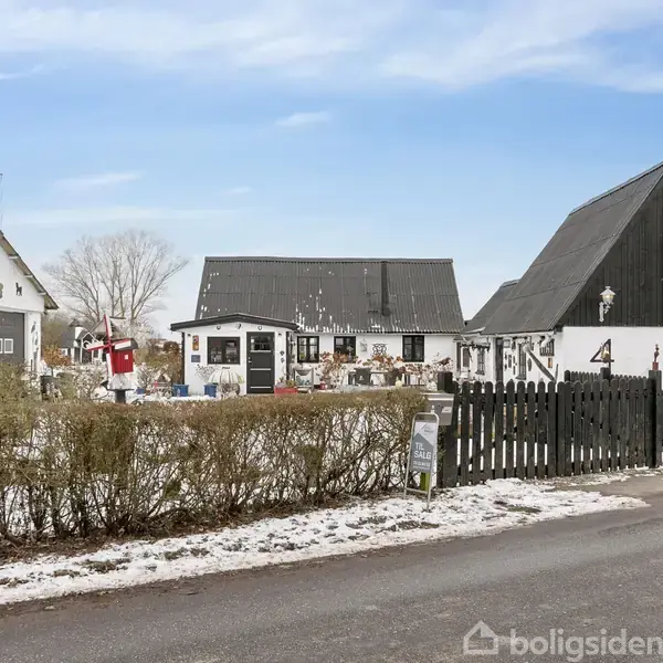 Hvide huse med sorte tage står i et snedækket område, omgivet af hække og en vej. Der er juledekorationer på husene og et sort hegn ved indgangen.