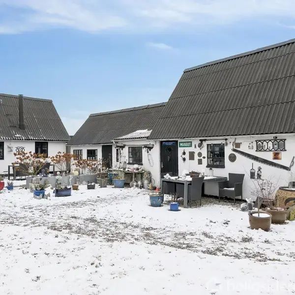 To hvide bygninger med sorte tage står på et snedækket gårdsplads, omgivet af potteplanter og sjove dekorationer. En dør har et skilt med teksten "ATELIER."