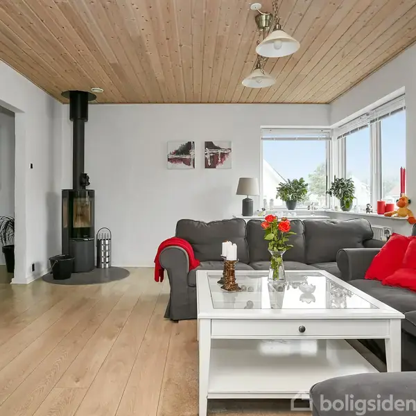 Stue med grå sofaer arrangeret omkring et hvidt sofabord med blomster. Trægulv og -loft. Vinduer med udsigt og en sort brændeovn i hjørnet. Vægur hænger til højre.