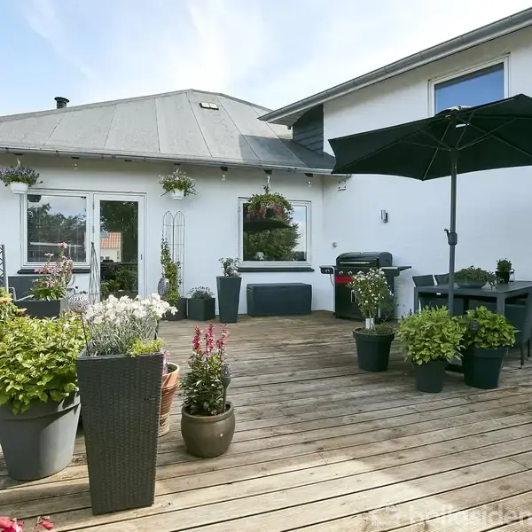 Terrasse med trædæk indeholder krukker med planter og blomster. Et sort parasol står ved siden af et spisebord. I baggrunden ses en grill og hvid bygning.