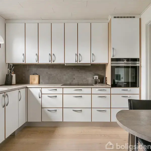 Et moderne køkken med hvide skabe og stålgreb. Bordet har frugter i en skål. Ovnen er indbygget i et skab. To lamper hænger over bordet.
