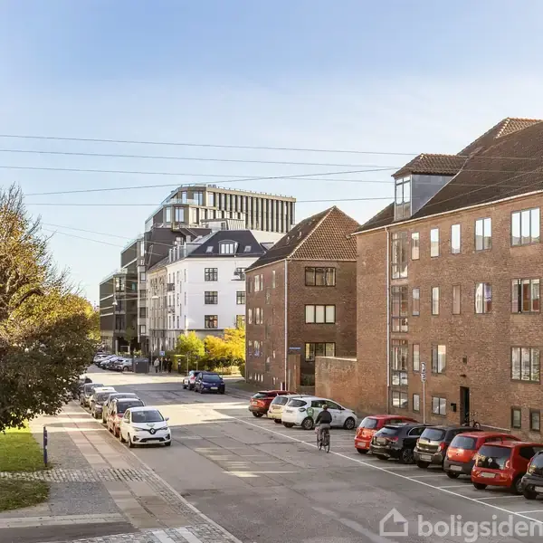 Rødstensbygninger står langs en byvej, hvor biler er parkeret. En person går på fortovet ved siden af træer under en klar blå himmel uden synlig tekst.