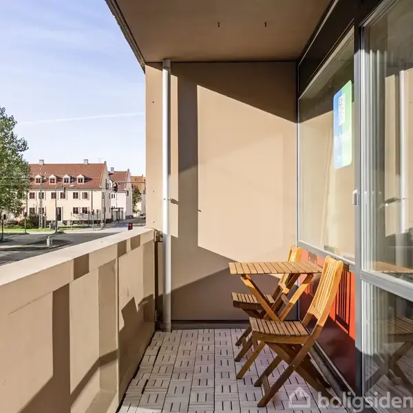Balkon med foldestole og bord vender mod gaden. Fliser på gulvet og bygninger samt træer i baggrunden skaber en byscene.