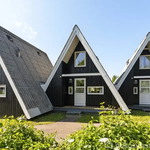 Tre sorte A-formede huse står stille, omgivet af grønne buske og en klar blå himmel i baggrunden.