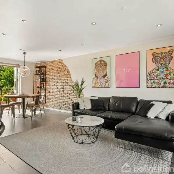 En stue med en sort lædersofa står mod en væg med tre kunstplakater. Et rundt sofabord står foran sofaen. Spisebord og stole står nær en åben dør mod en terrasse. En murstensvæg tilføjer karakter.