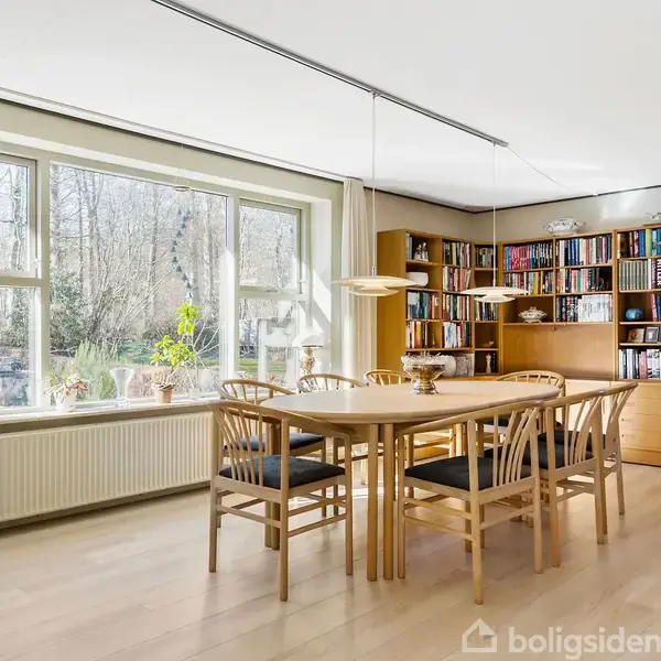 Stue med spisebord og stole, omgivet af store vinduer der giver udsigt til haven. En bogreol står ved siden af et vitrineskab. Et sofabord er pyntet med en skål og dug.