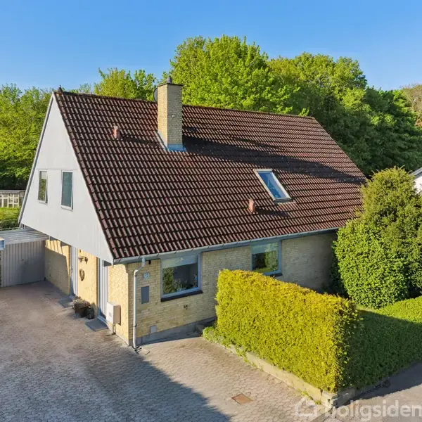 Et to-etagers hus står på en flisebelagt indkørsel, med en carport til venstre. Det har en brun tagstenstag, en gul murstensfacade og er omgivet af grønne buske og træer.