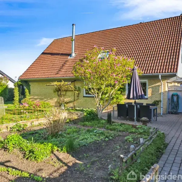 Huset med rødt tag står i baggrunden, omkranset af en have med bede og en træterrasse. En spiseplads med parasol står ved siden af en brolagt sti.