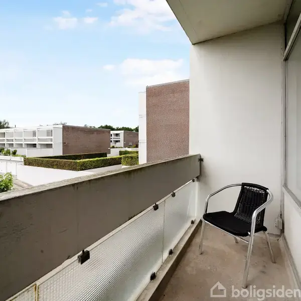 En sort stol står stille på en altan med glasværn. Omgivelserne viser et boligkompleks med flade tage og grønne buske i det fjerne under en klar himmel.