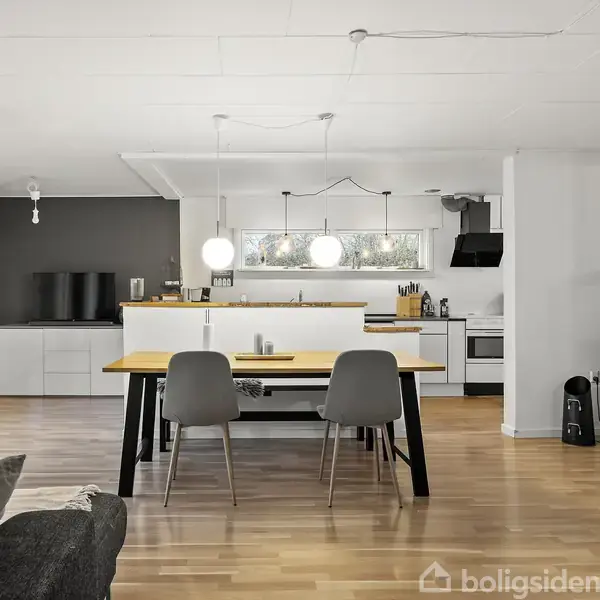 Et åbent køkken-alrum med spisebord og stole centreret, omgivet af moderne køkkenudstyr og en brændeovn på trægulv. Store vinduer giver lys, og en tv-skærm ses i baggrunden.