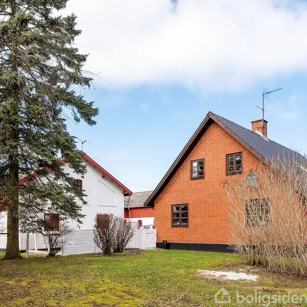 Et rødt murstenshus med sort tag står ved siden af et hvidt hus i en have. En høj grantræ står foran dem under en delvis skyet himmel.
