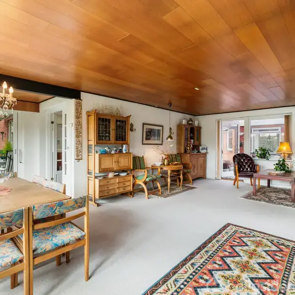 En stue med træloft har en spisebord med stole og en lysekrone. Et tæppe med mønster ligger på gulvet. I baggrunden ses stole og et sofabord nær en dør til haven.