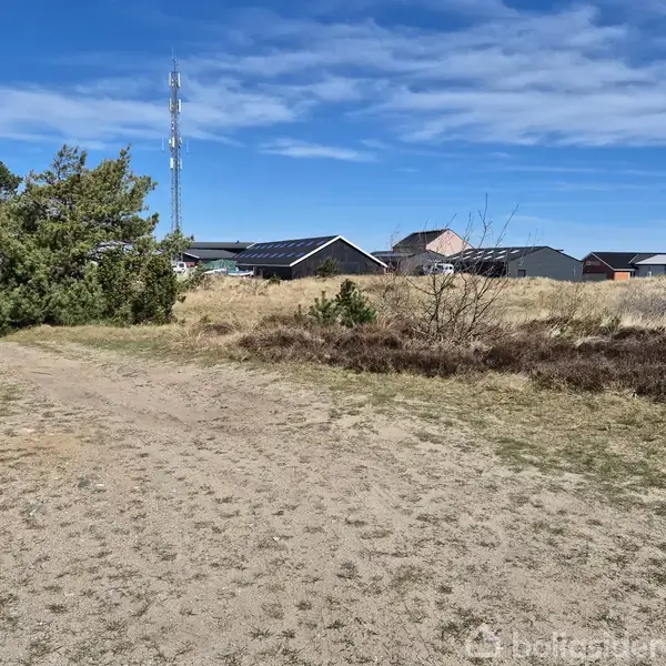 Sti i sandet; snoer sig gennem tørt landskab med buske og træer; i baggrunden ses huse og en mobilmast under en blå himmel med lette skyer.
