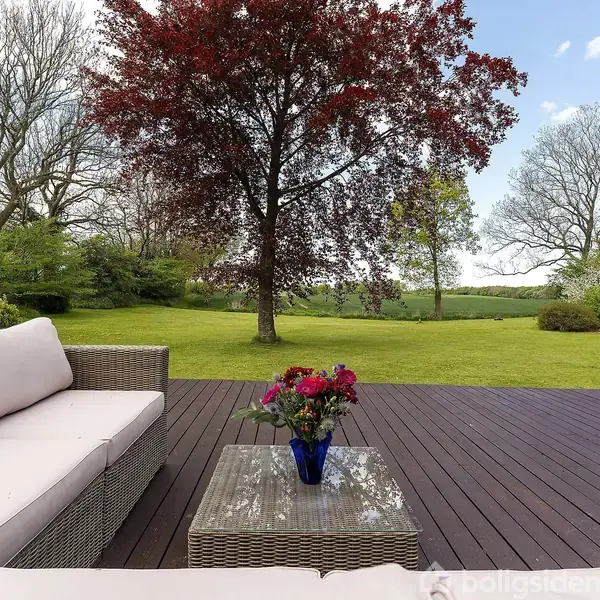 Sofa og bord med blomster på en terrasse. Træ i baggrunden omgivet af frodig græsplæne og natur. Klar himmel skaber en afslappende udendørs atmosfære.