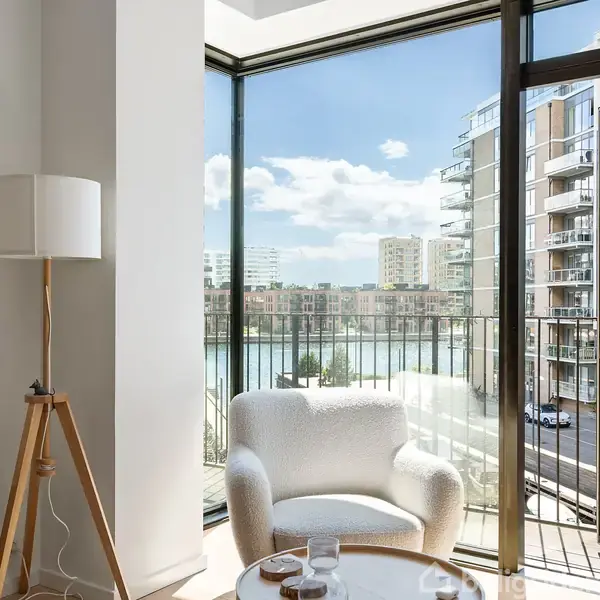 En hvid lænestol står foran en stor glasdør, der åbner ud til en altan med udsigt over moderne lejlighedsbygninger og vand. En gulvlampe er placeret ved siden af stolen.