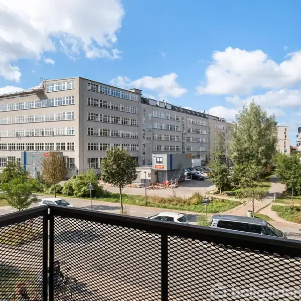 Luftigt bybillede. Moderne bygninger omkranser en vej med parkerede biler og frodige træer. Klar blå himmel og skyer set fra en balkon med sort gitter.
