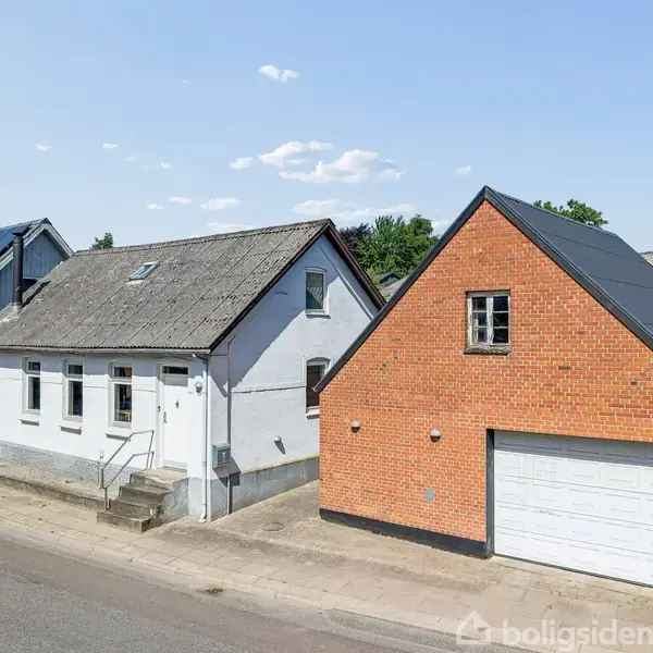 Et hvidt hus med en lille trappe ved indgangen står ved siden af en rød murstensbygning med en garage. Omgivet af en vej og grønne træer under en blå himmel.