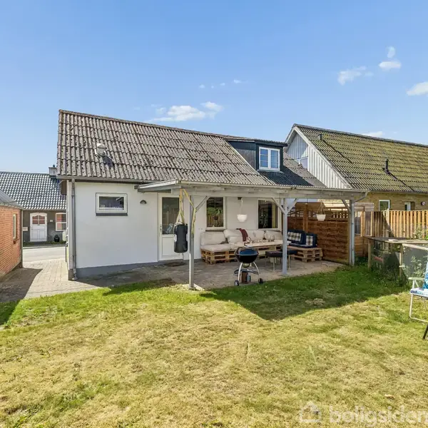 Et enfamilieshus med græsplæne, overdækket terrasse og grill. Huset har hvid facade og skråt tag. Omkranset af nabohuse og træhegn, under en skyfri himmel.