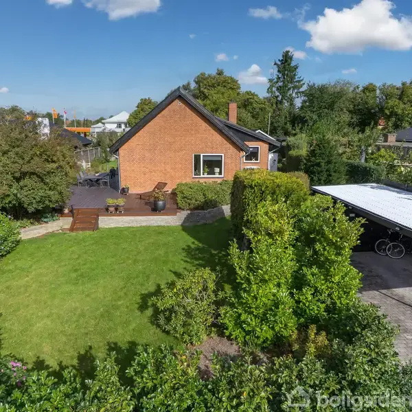 Rødt murstenshus står i en frodig have med græsplæne og træer. Der er en terrasse med havemøbler og en carport til højre under en klar blå himmel.