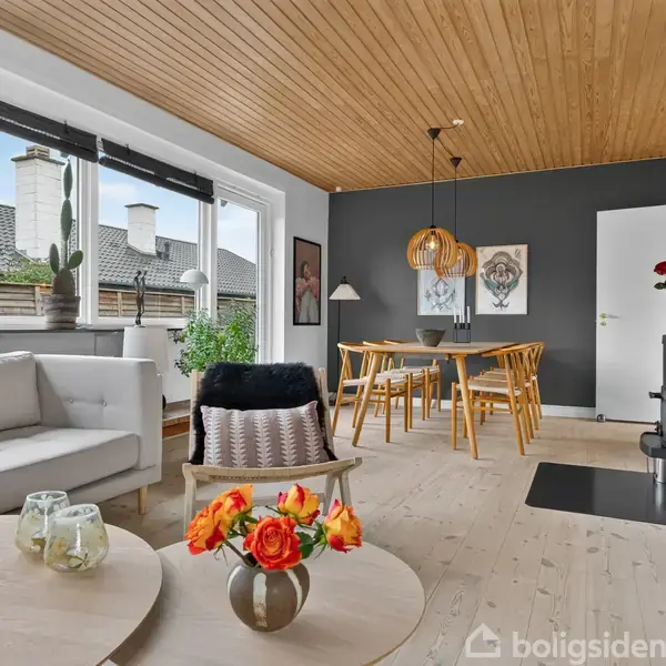 Stue med lyse trægulve og træloft, indeholder sofa, rundt bord med blomster, spisebord med stole. Store vinduer giver udsigt til hustage. Moderne indretning med kombineret opholds- og spisestue.