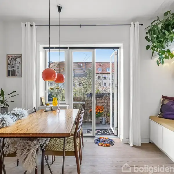 Spisebord med stole er placeret ved store, åbne døre til en altan. Rummet har lyse gardiner, hængende planter og en hylde med bøger. Lyse nuancer og enkel indretning dominerer.