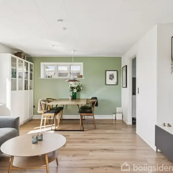 Stue med grå sofa og rundt sofabord. Spisebord med seks stole står foran en lysegrøn væg. TV og reolsystem fylder højre side af rummet.