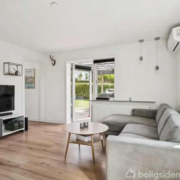 Stue med grå hjørnesofa, fladskærms-TV på lavt møbel. Trægulve, hvide vægge. Store vinduer og udgang til terrasse. Hylder med planter og dekorationer. Airconditionenhed på væggen.