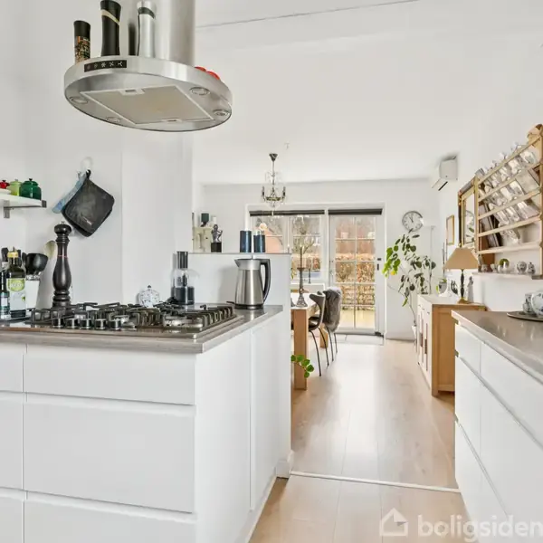 Køkken med moderne hvide skabe og en kogezone med emhætte, fører til en stue med spisebord. Lyse trægulve og vinduer giver et lyst indtryk.
