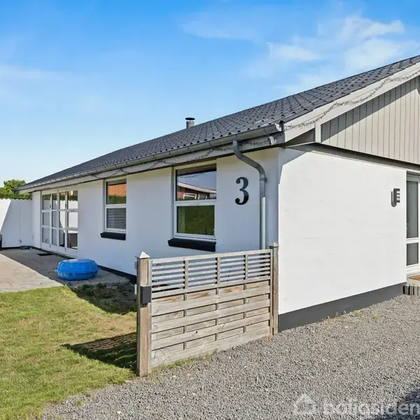 Et hvidt, enkeltstående hus med nummer 3 på facaden står på en græsplæne ved siden af en terrasse med bord og stole, omgivet af hegn.