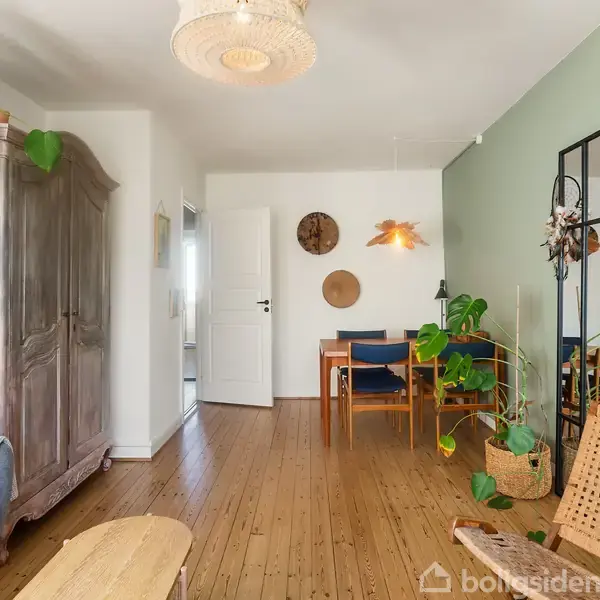 Et træskab står i en stue med en grøn væg, hvor planter og møbler skaber en hyggelig atmosfære. Et spisebord med stole ses i baggrunden.