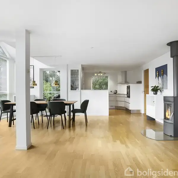 Stue med brændeovn i hjørnet, stilfuldt spisebord og stole. Store vinduer giver udsigt til frodigt udendørsområde. Moderne køkken i baggrunden, træparketgulve og lyse vægge. Ingen tekst fundet.