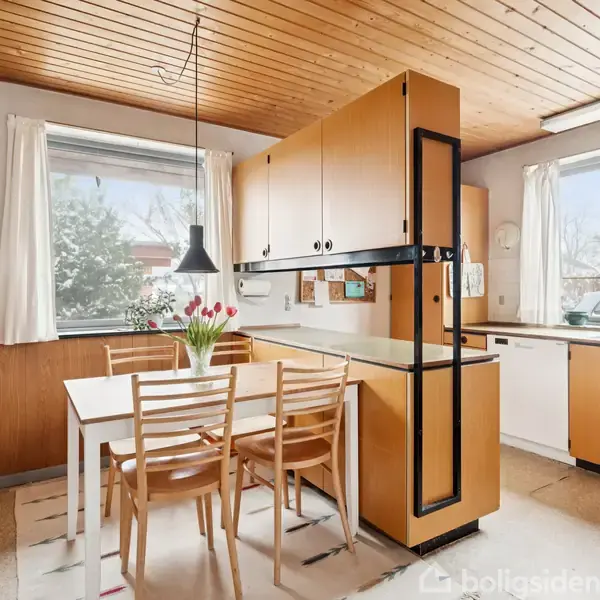 Et køkken med træloft indeholder et spisebord med fire stole, en vase med tulipaner og hængelampe. Skabe i lysebrun rammer køkkenet ind, og store vinduer slipper lys ind.