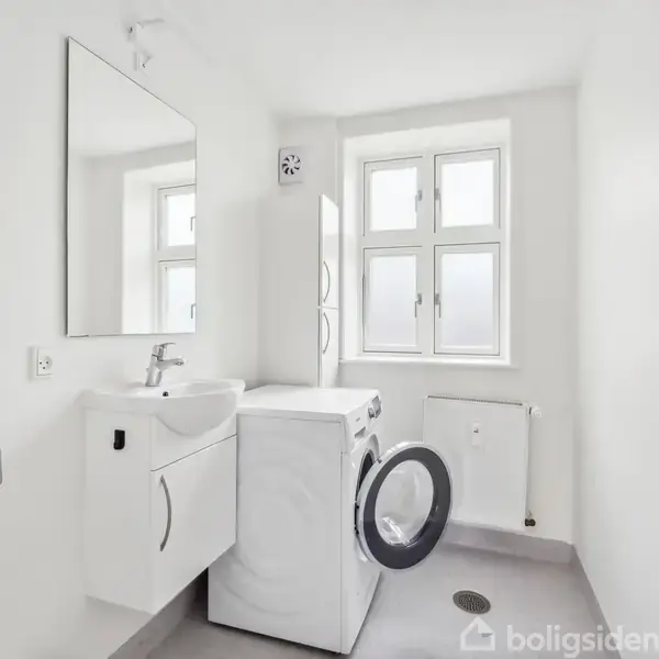 Vaskemaskine med åben dør står ved siden af hvidt badeværelsesskab og vask. Hvidt rum med spejl, to vinduer og radiator. Dør til højre. Ingen synlig tekst udover "Boligsiden".