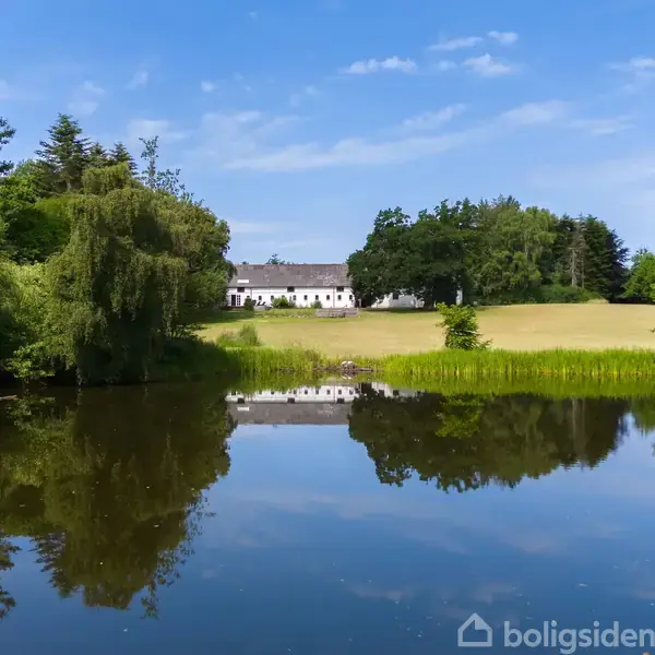 En hvid bygning reflekteret i en rolig sø, omgivet af frodig, grøn vegetation og marker under en klar blå himmel.