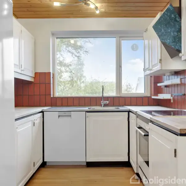 Køkken med hvide skabe og apparater, herunder opvaskemaskine og ovn, står stille. Røde flisevægge matcher træloft. Stort vindue giver udsigt til træer. Ingen tekst fundet.