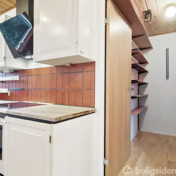 Et køkken med hvide skabe og en ovn står i forgrunden. Orange fliser pryder væggen. Til højre er en gang med flere åbne hylder på væggen.