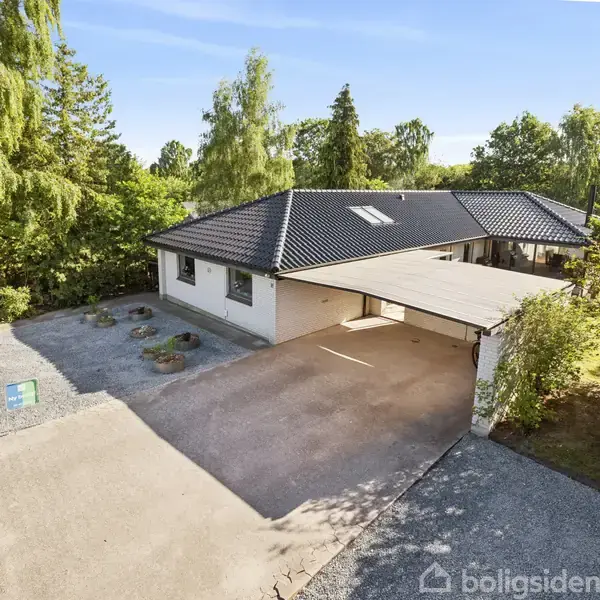 Et hvidt hus med sort tag står stille med tilhørende carport. Omkranset af grønne træer og buske, huset ligger langs en stille vej med grusindkørsel. Skilt er ved vejen.