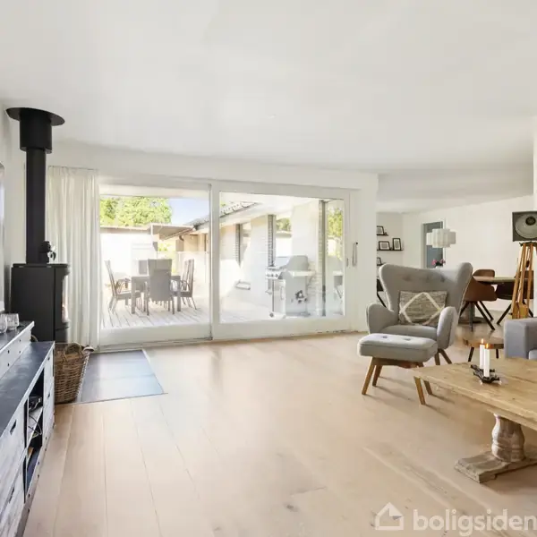 Stue med brændeovn og stort vindue mod terrasse. Lys indretning med trægulv, sofa, lænestol og reol. Terrassebord ses udenfor gennem glasset. Stemning af moderne hygge.